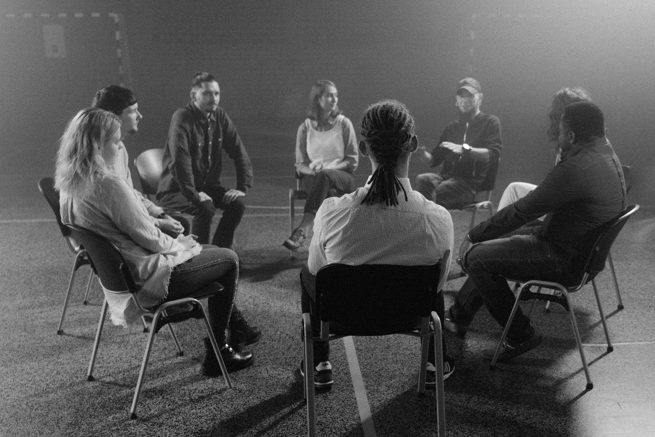 Home A diverse group of people in a circle discussing and supporting each other in a therapy session.